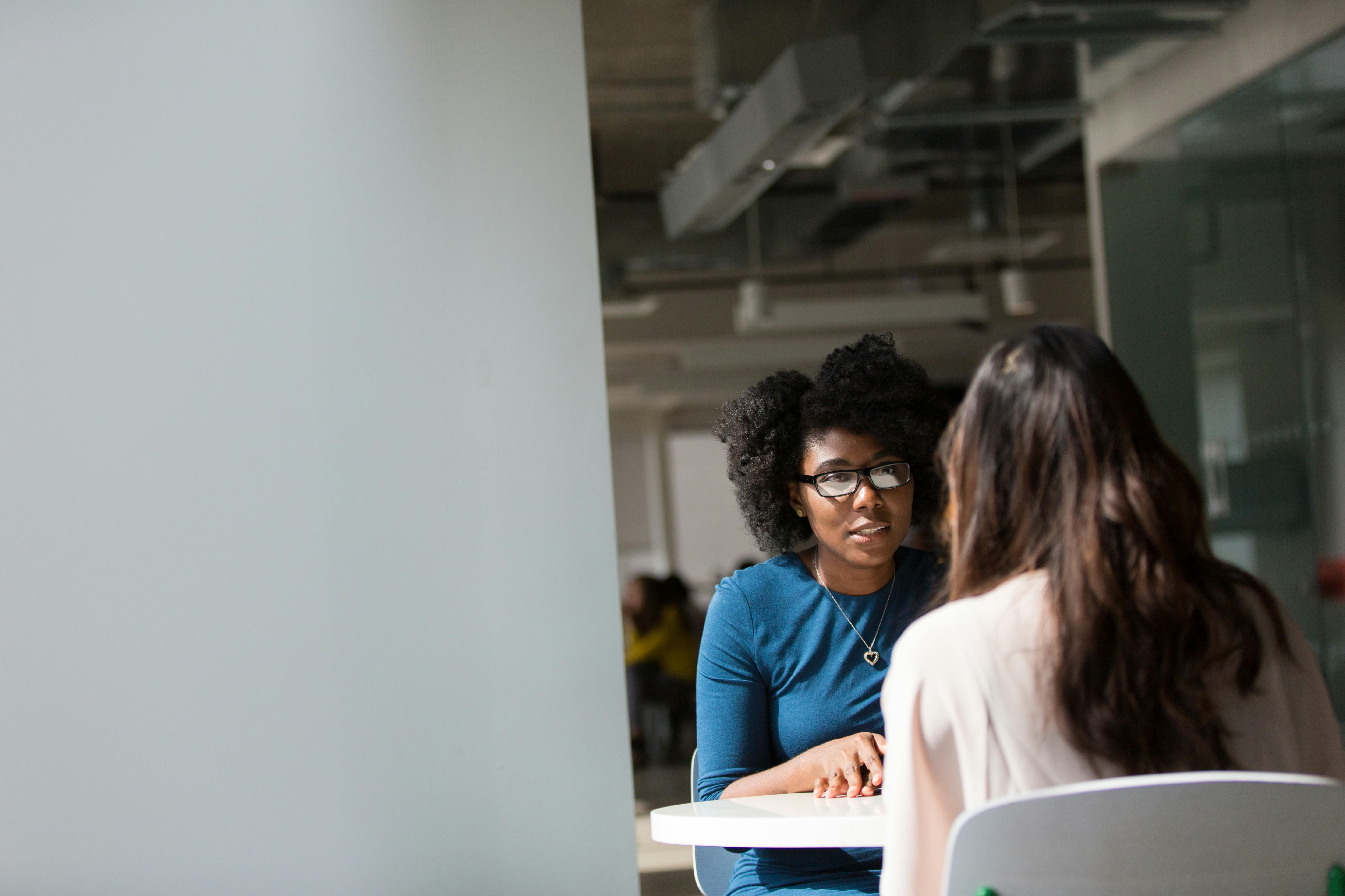 A woman with curly hair and glasses is engaged in a conversation with another woman at a table in a well-lit office environment.